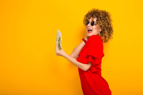 Щаслива молода кучерява жінка в червоній сукні та сонцезахисних окулярах тримає гроші
  - Фото, зображення