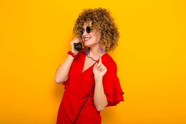 Atraktivní mladá kudrnaté vlasy žena v červených šatech a sluneční brýle, mluví na vinobraní drátu telefonní  - Fotografie, Obrázek