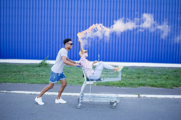 Young hipsters couple riding on trolley on empty shop parking during sunrise and have good funny time - 写真・画像