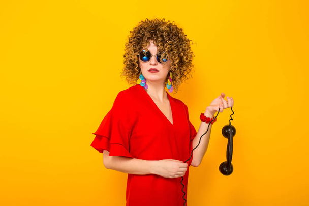 Attractive young curly haired woman in red dress and sunglasses holding handle of vintage phone - Photo, Image