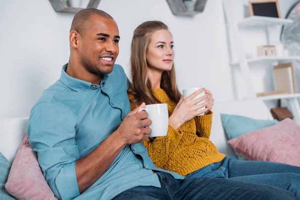 multicultural couple sitting on sofa with coffee and looking away - Φωτογραφία, εικόνα
