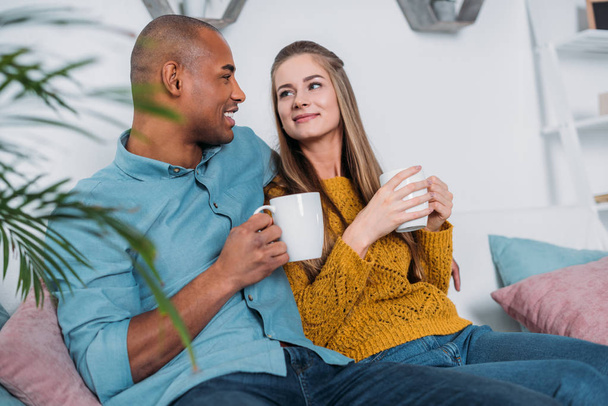 smiling multicultural couple sitting on sofa with coffee - Foto, afbeelding