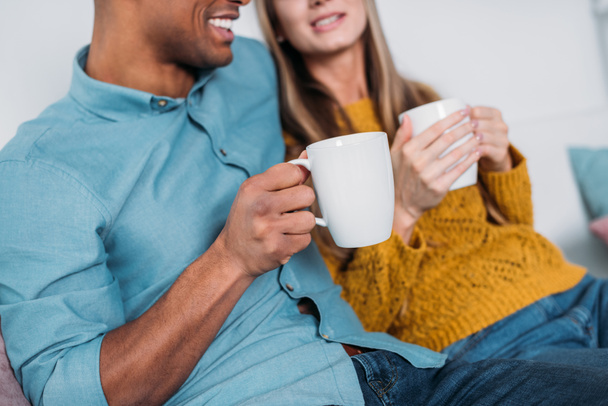cropped image of smiling multicultural couple sitting on sofa with coffee - Fotó, kép
