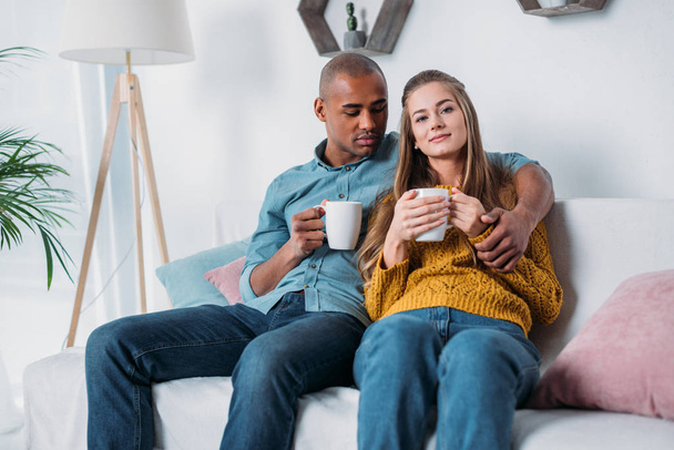pareja multicultural abrazos y sentarse en el sofá con café
 - Foto, Imagen