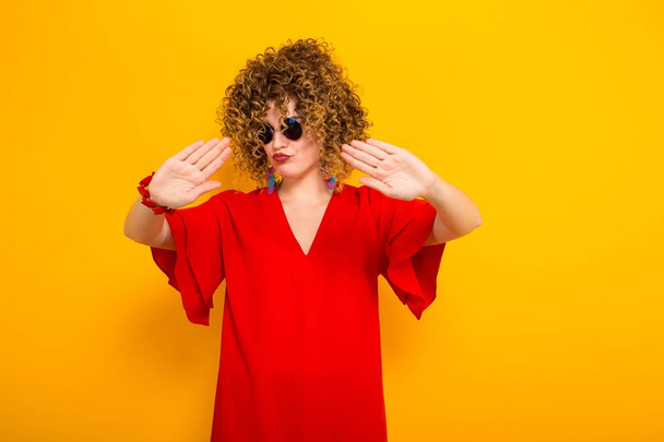 Attractive young curly haired woman in red dress and sunglasses showing stop gesture - Foto, afbeelding
