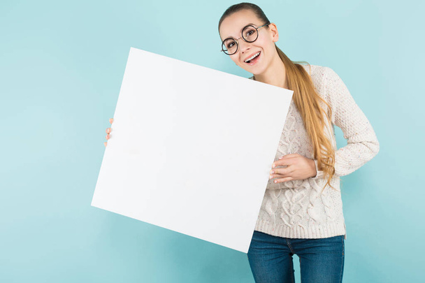 Hezká dívka v stylové brýle drží prázdný nápis modré studio pozadí  - Fotografie, Obrázek