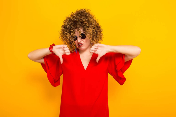 Attractive young curly haired woman in red dress and sunglasses showing thumbs down - Foto, immagini