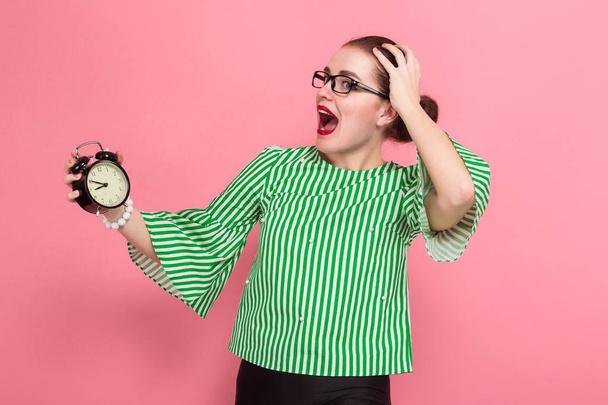 Shocked beautiful young woman in stylish eyeglasses holding alarm clock on pink background  - Photo, Image