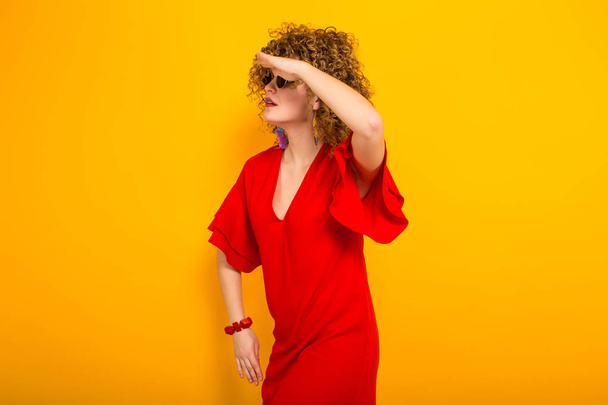 Attraente giovane donna dai capelli ricci in abito rosso e occhiali da sole alla ricerca di qualcosa
  - Foto, immagini