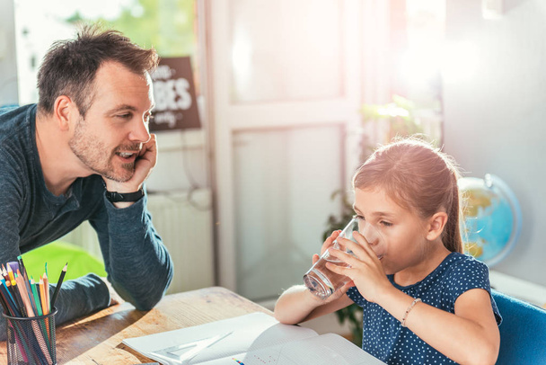 Girl drinking water while doing homework and father standing next to her - Photo, Image