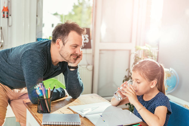 Girl drinking water while doing homework and father standing next to her - Foto, Bild