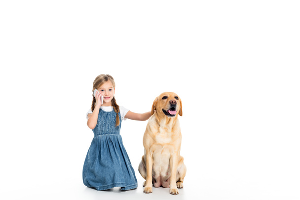 adorable kid sitting with dog while talking on smartphone, isolated on white - Foto, Imagen
