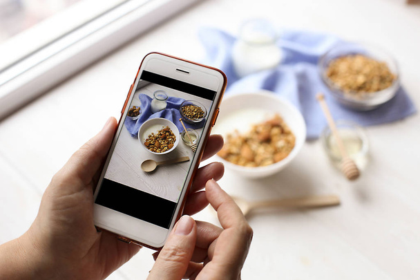 Close up of female hands holding mobile phone taking a picture of granola cereals, milk yogurt and trail mix vegetarian dieting breakfast for a new food blog article. Top view, overhead, copy space. - Φωτογραφία, εικόνα