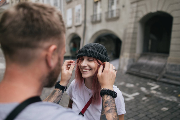 επιλεκτική εστίαση του ανθρώπου βάζοντας καπέλο σε όμορφη χαμογελαστή νεαρή γυναίκα ενώ περπατούν μαζί στην οδό στη Βέρνη, Ελβετία  - Φωτογραφία, εικόνα