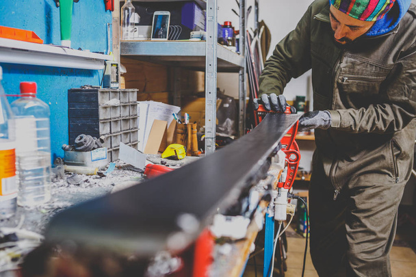 Um trabalhador masculino em uma oficina de serviço de esqui repara a superfície de deslizamento dos esquis. Close-up de uma mão com um raspador de plástico para remover a cera, removendo a nova cera. Reparação de tema de freio de esqui
 - Foto, Imagem