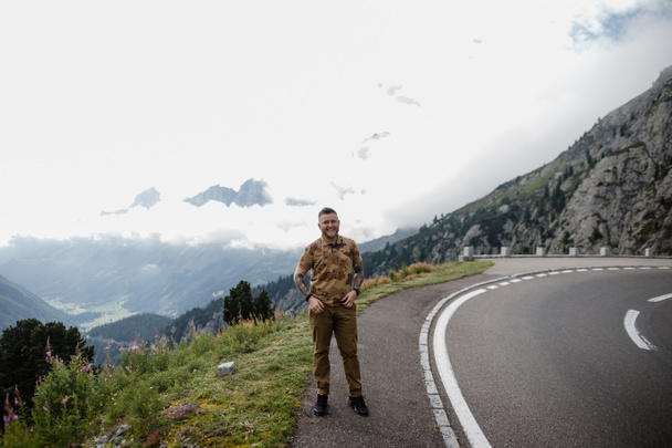 joven guapo de pie en el camino de asfalto en las montañas y sonriendo a la cámara, Alpes
  - Foto, Imagen