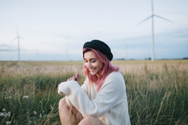bonita chica con pelo rosa en suéter blanco y sombrero sentado en el campo con molinos de viento
 - Foto, Imagen
