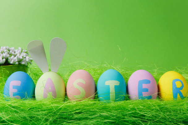 Fila de seis coloridos huevos de Pascua pintados monofónicos pastel con inscripción Pascua, divertidas orejas de conejo, flores blancas en cubo en hierba verde aislada sobre fondo verde. Copiar espacio para publicidad
. - Foto, imagen