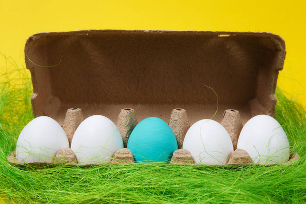 Sorban a négy fehér, egy pasztell egyszólamú türkiz festett húsvéti tojás karton tálca sárga háttéren zöld fűben. Boldog húsvéti koncepció. Másolja a hirdetés terület. A hely a szöveg. - Fotó, kép