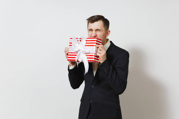 junger gutaussehender lustiger Mann im Anzug beißt roten Geschenkkarton mit Geschenk isoliert auf weißem Hintergrund. Kopierfläche für Werbung. st. valentinstag, internationaler frauentag, geburtstag, ferienkonzept. - Foto, Bild