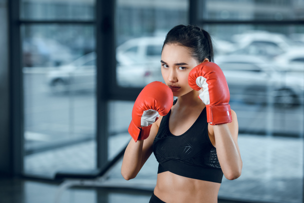 jonge vrouwelijke bokser in handschoenen bij sportschool kijken camera - Foto, afbeelding