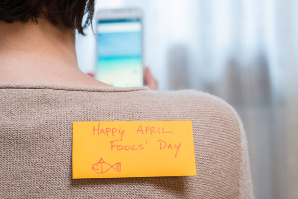 Feliz Dia dos Tolos de Abril. Mulher usando um telefone celular com uma nota pegajosa em suas costas
 - Foto, Imagem