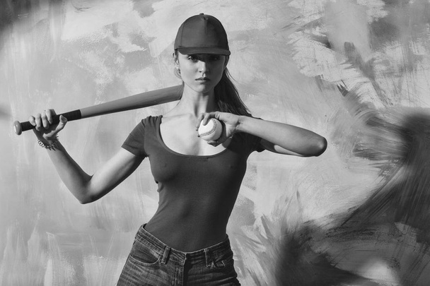 Pretty girl or sexy baseball player with blond hair in red cap, tshirt and blue jeans standing with green bat and ball on abstract yellow and pink wall - Fotografie, Obrázek