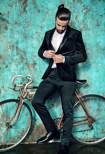 portrait of handsome fashion stylish hipster lumbersexual businessman model dressed in elegant black suit posing near sport bicycle and blue grange wall in studio. Metrosexual - Foto, immagini