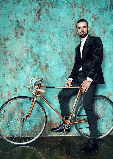 portrait of handsome fashion stylish hipster lumbersexual businessman model dressed in elegant black suit posing near sport bicycle and blue grange wall in studio. Metrosexual - Foto, Imagem