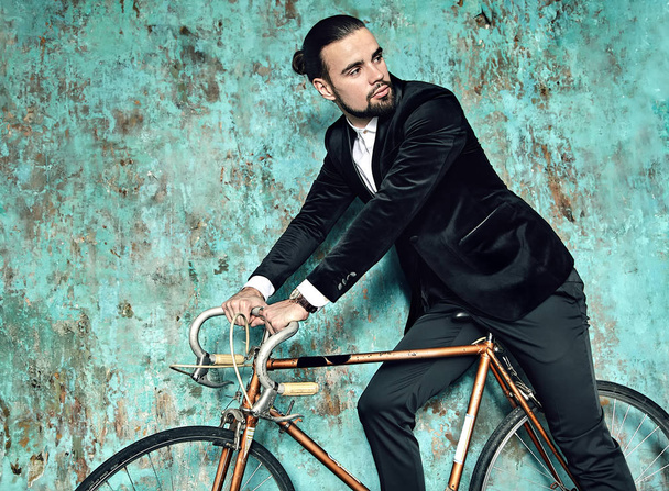 portrait of handsome fashion stylish hipster lumbersexual businessman model dressed in elegant black suit posing near sport bicycle and blue grange wall in studio. Metrosexual - Fotó, kép