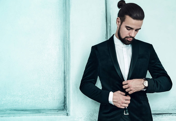 portrait of handsome fashion stylish hipster lumbersexual businessman model dressed in elegant black suit in posing near light blue wall in studio. Metrosexual - Foto, Bild