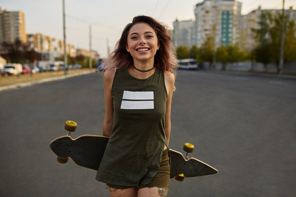 Όμορφο κορίτσι με τατουάζ που ποζάρει με ένα πολύχρωμο longboard - Φωτογραφία, εικόνα