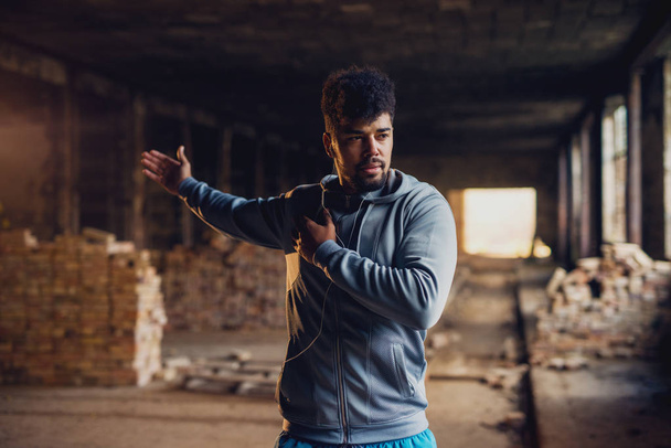 portrait of motivated afro-american man stretching in abandoned hangar place with earphones - Фото, зображення