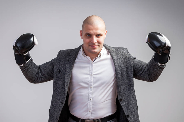 Young bald man in white shirt, gray suit and boxing gloves smiling, showing biceps on white isolated background - Photo, Image