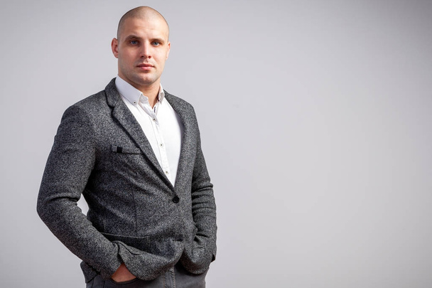 A young bald man in a white shirt, gray suit strictly looks into the camera and holds his hands in the pockets of his jacket on a white isolated background - Photo, Image