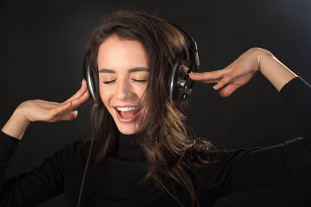 Эмоционально громко поющая женщина слушает музыку в беспроводных наушниках и рукой показывает знак v на темном фоне
 - Фото, изображение
