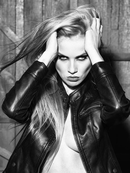 Portrait of one attractive sensual sexy young serious passionate blonde woman touching long hair with hands in leather black jacket in studio on wooden wall background, vertical picture - Photo, Image