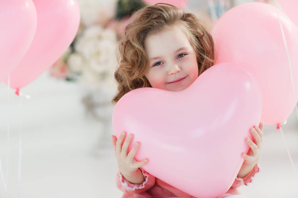 A little girl of 6 years with long curly hair, dressed in a pink dress and white tights, a beautiful smile, sits alone in a large bright room with lots of pink balloons in the shape of a heart. Valentine's Day and party celebration. - Foto, afbeelding
