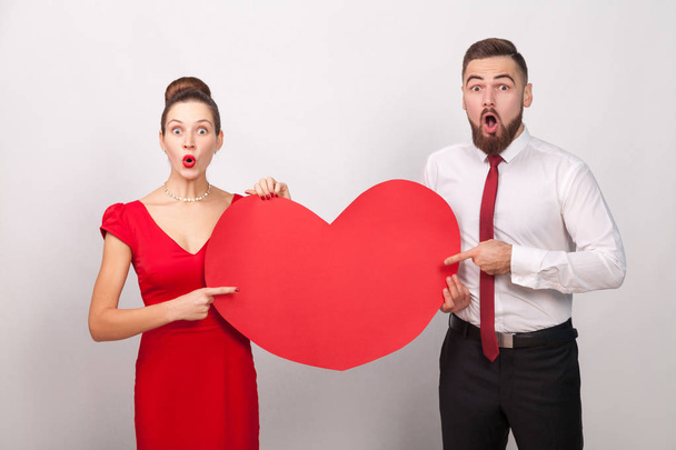 wondered couple with opened mouths pointing fingers at red heard on gray background, valentines day concept  - Photo, image