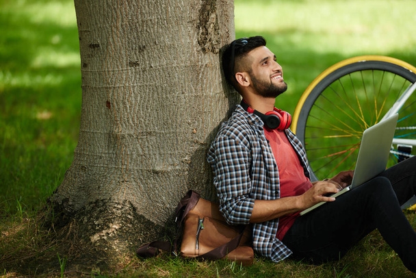 Улыбающийся молодой человек сидит под деревом с ноутбуком на коленях и смотрит вверх
 - Фото, изображение