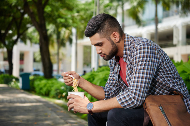 Jovem indiano sentado ao ar livre e comendo sopa de macarrão instantâneo para o almoço
 - Foto, Imagem