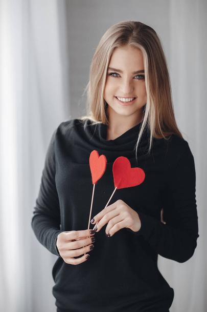 Young beautiful woman with a nice smile and big grey eyes,a blonde with long straight hair,dressed in a black party dress, spends time alone in the big bright room,holding hands of little Krasnye the heart is a symbol of Valentine's day - Foto, imagen