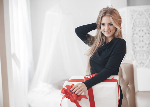 A young beautiful woman with a pleasant smile and big gray eyes, a blonde with long straight hair dressed in a black festive dress, alone in a large bright room examines gifts in white boxes for Valentine's Day - Foto, imagen