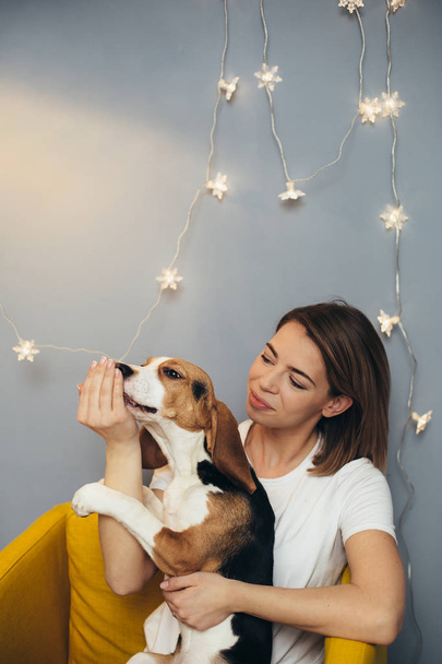 Young woman in white t-shirt hug beagle puppy on yellow chair with grey wall and on background - Foto, Imagen