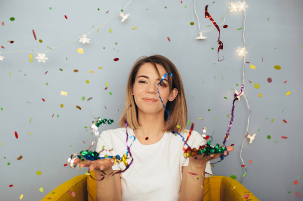 Young woman in white t-shirt blast confetti on yellow chair with grey wall on background - Фото, зображення