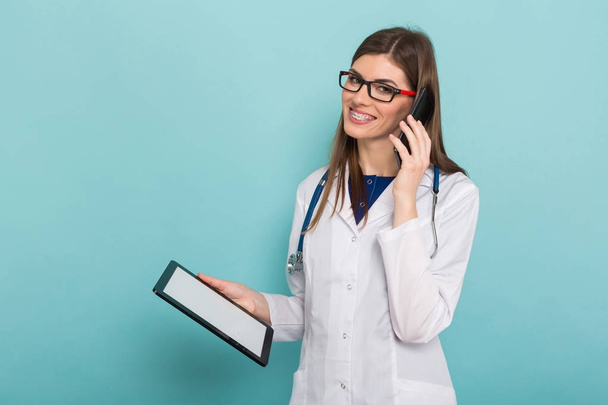Dijital tablet tutarak ve smartphone üzerinde konuşurken stetoskop ile kendine güvenen genç bir kadın doktor - Fotoğraf, Görsel