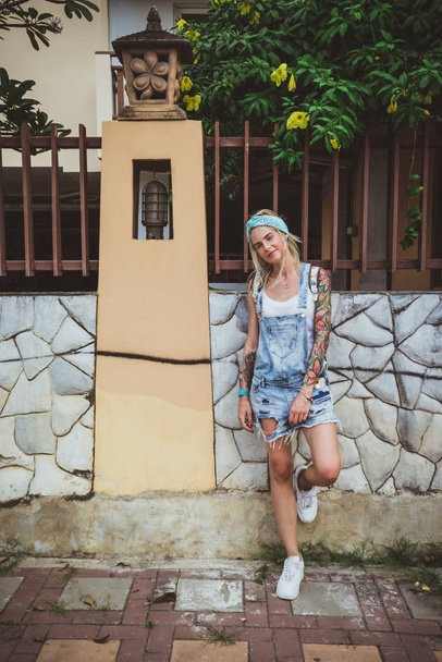 Beautiful girl posing near a fence. He wears a denim overall and bandana. A modern student. Tattoo model - Foto, Imagen
