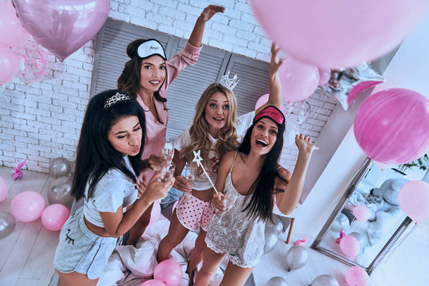 Vista superior de cuatro mujeres sonrientes jóvenes atractivas que tienen fiesta de pijamas en el dormitorio con alcohol y globos
 - Foto, Imagen