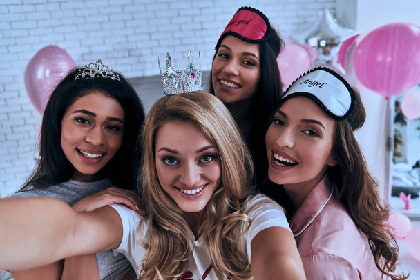 Self portrait of four playful young women smiling and looking at camera while having a slumber party, wearing diadem, princess crown and sleep masks - Foto, immagini