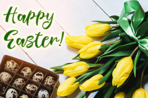 Easter spring decorative composition. Bouquet of yellow tulips tied by green ribbon and little box with quail eggs. Close up portrait on white wooden background. - Photo, image